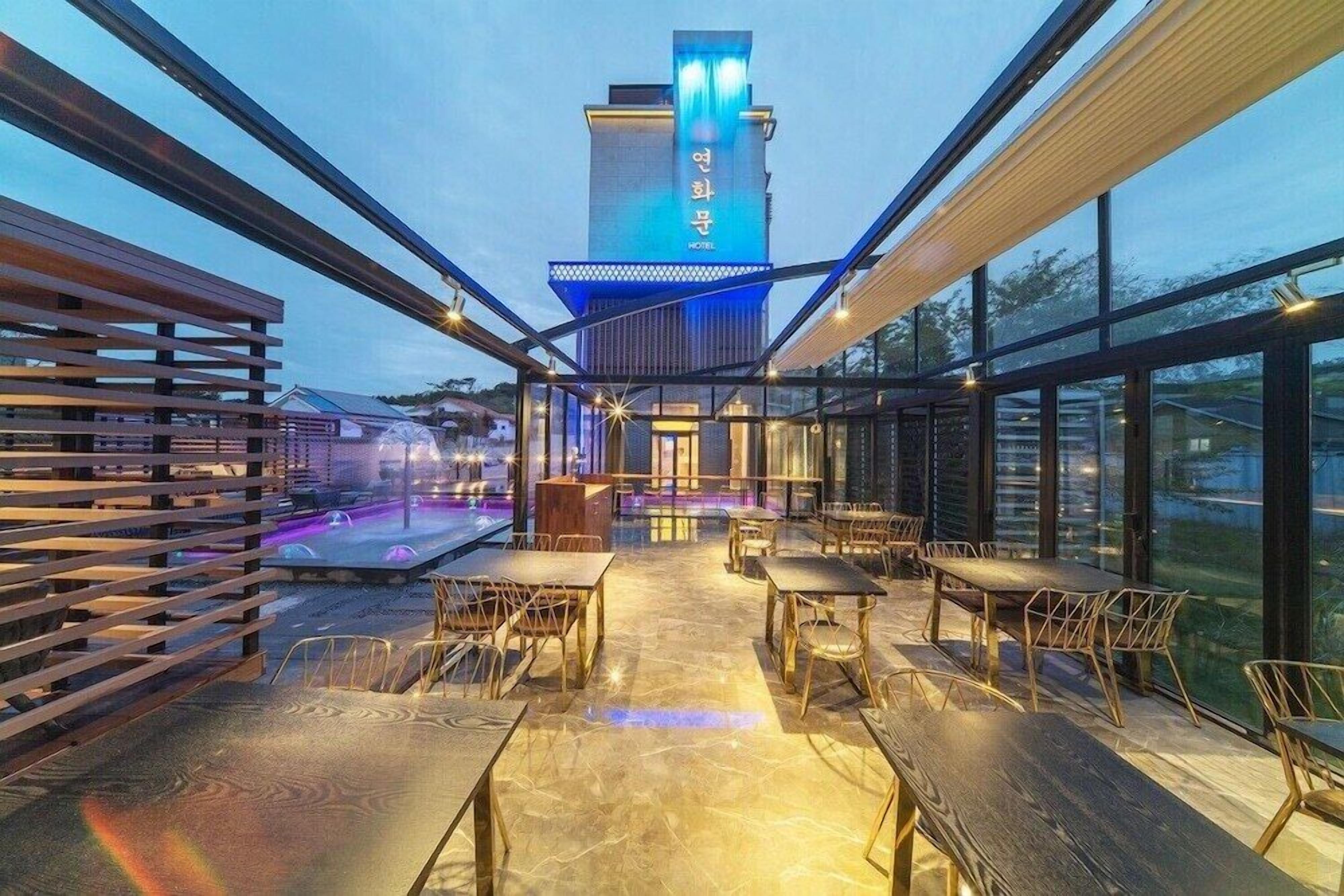 Un área de comedor al aire libre con mesas y sillas, una piscina y un edificio con señalización iluminada al fondo.