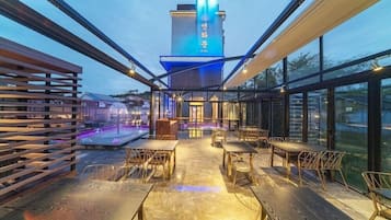 Un área de comedor al aire libre con mesas y sillas, una piscina y un edificio con señalización iluminada al fondo.