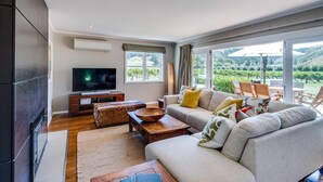 A modern living room with a flat-screen TV, a sofa, a coffee table, and a view of a vineyard through large windows. - The Farmhouse At Mission Estate (Napier)