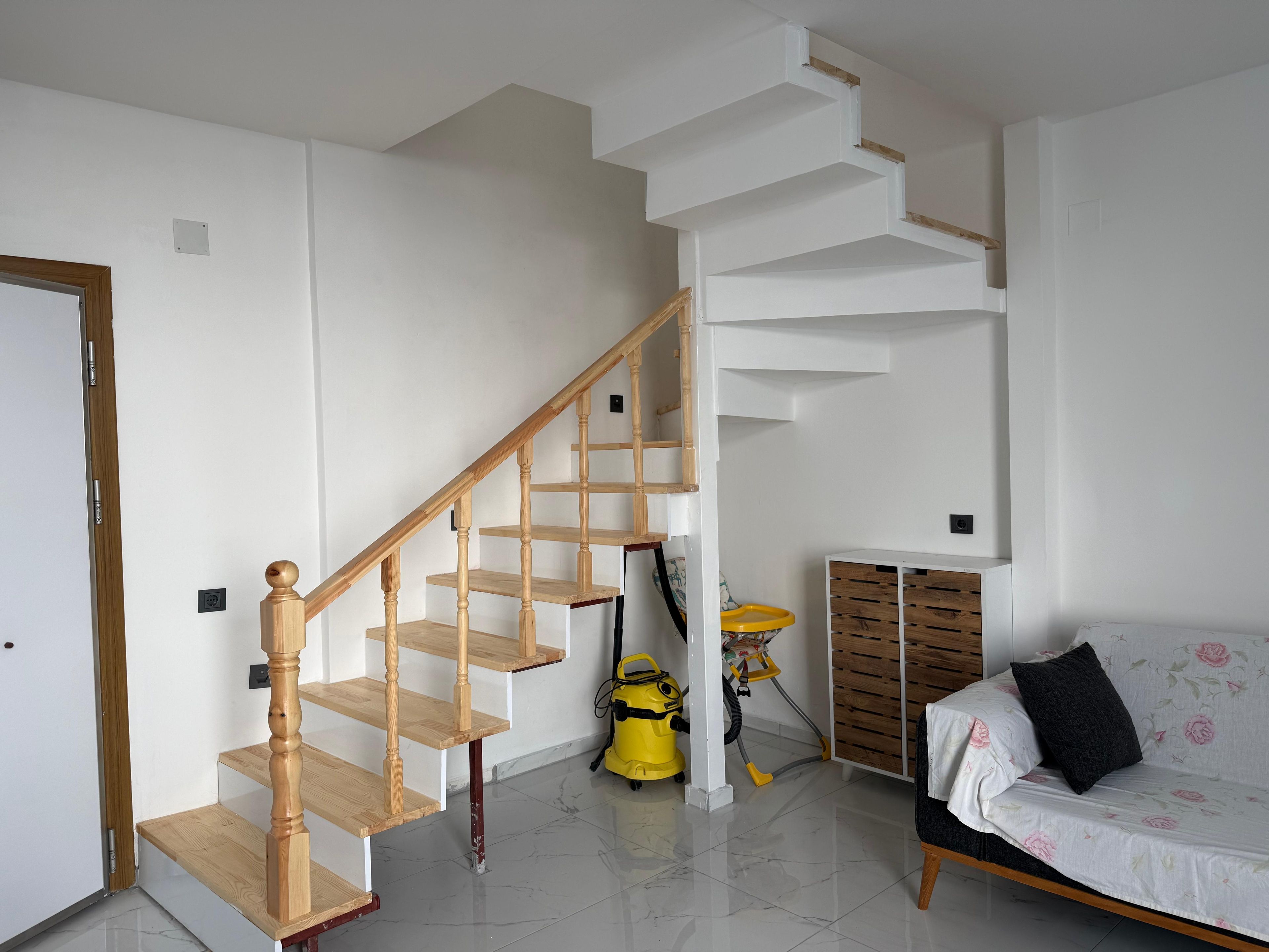 Eine Treppe mit hölzernen Geländern, ein gelber Staubsauger, ein Sofa mit Blumenmuster und ein Holzschrank.