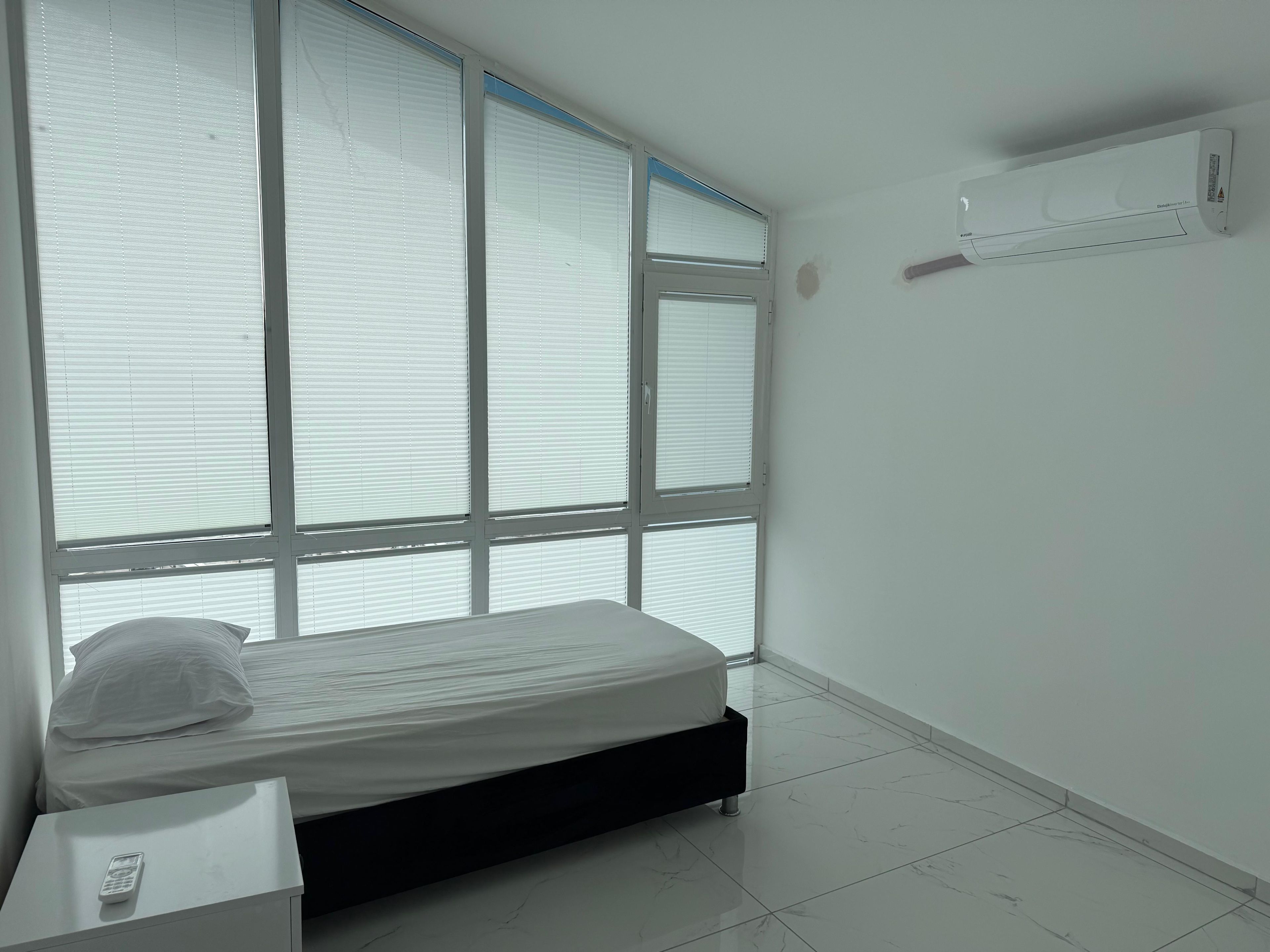 Ein Schlafzimmer mit einem Bett, einem Nachttisch, einer Fernbedienung und einem Fenster mit Jalousien.