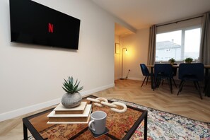A modern living room with a flat-screen TV, a dining area with chairs and a table, and a large window with a city view.