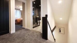 Un couloir au sol recouvert de moquette, un escalier avec une rampe noire et une chambre à coucher visible à travers une porte ouverte.