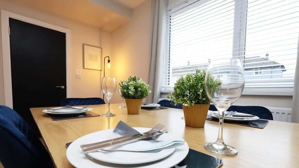 Une table à manger dressée pour deux personnes, avec assiettes, couverts et verres à vin, encadrée de plantes en pot, dans une pièce dotée d’une fenêtre et d’une porte.