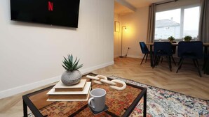 Un salon moderne avec un téléviseur à écran plat, une table basse avec des livres et une plante, un coin repas avec des chaises et une table, et une grande fenêtre avec des rideaux.