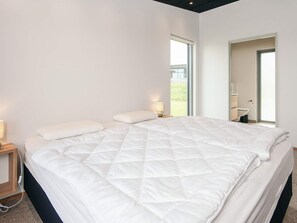 Una cama grande con edredón blanco acolchado, una mesita de noche con lámpara y una ventana con vistas a la vegetación.