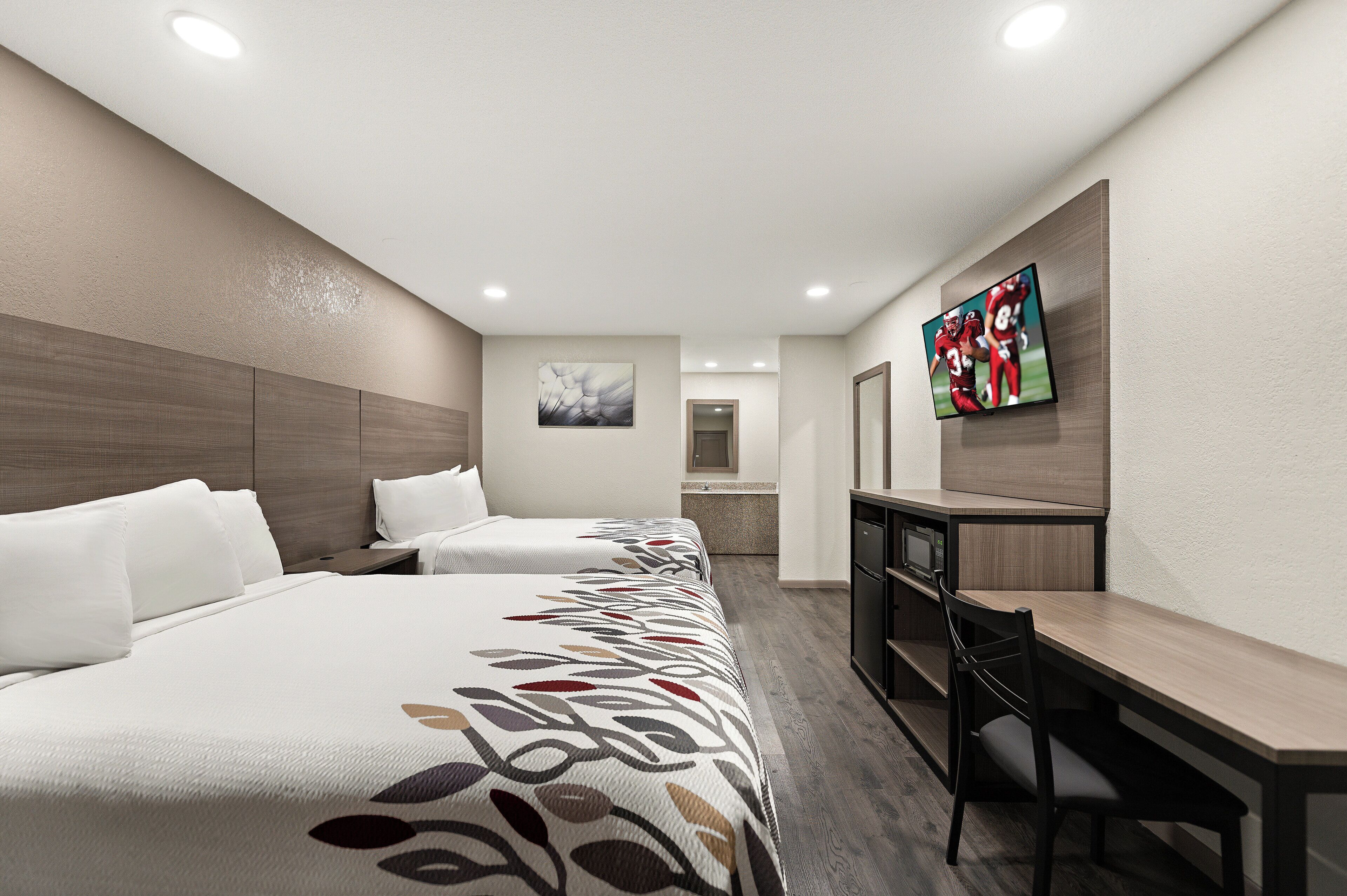 A hotel room with two beds, a desk, a television, and a bathroom in the background.
