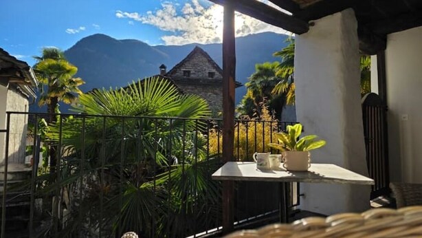 A balcony with a view of mountains, a rustic building, and tropical plants. - Cavigliano Rustic (Cavigliano)