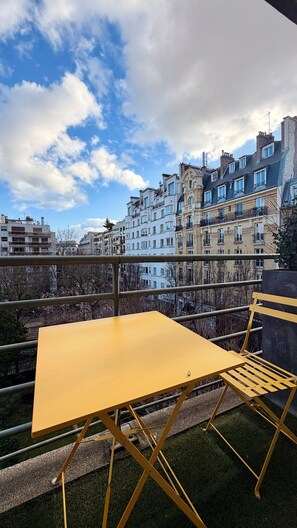 Et gult bord og en stol på en balkon med udsigt til høje bygninger og et delvist overskyet himmelbillede.