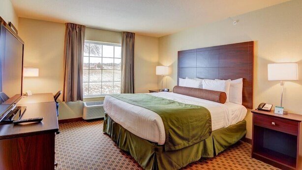A hotel room with a large bed, a desk, a chair, a telephone, and a window with curtains. - Cobblestone Hotel Suites Punxsutawney Pa (Punxsutawney)