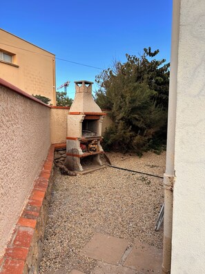 A small outdoor brick oven structure with a chimney, surrounded by gravel and a low brick wall. - Ground-floor apartment in a villa overlooking the beach (Valras-Plage)