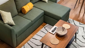 A green sectional sofa with a rolled-up blanket and patterned pillows, a wooden coffee table with a remote control and woven placemats, and a wooden chair. - Brand-new Fulham Stay Near Thames (London)