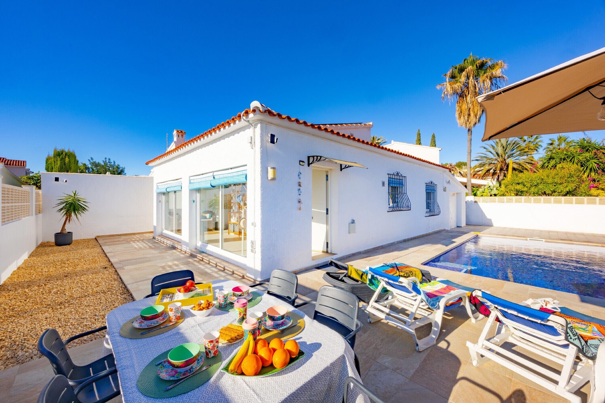A white building with a tiled roof, a swimming pool, and outdoor seating with tables and chairs.