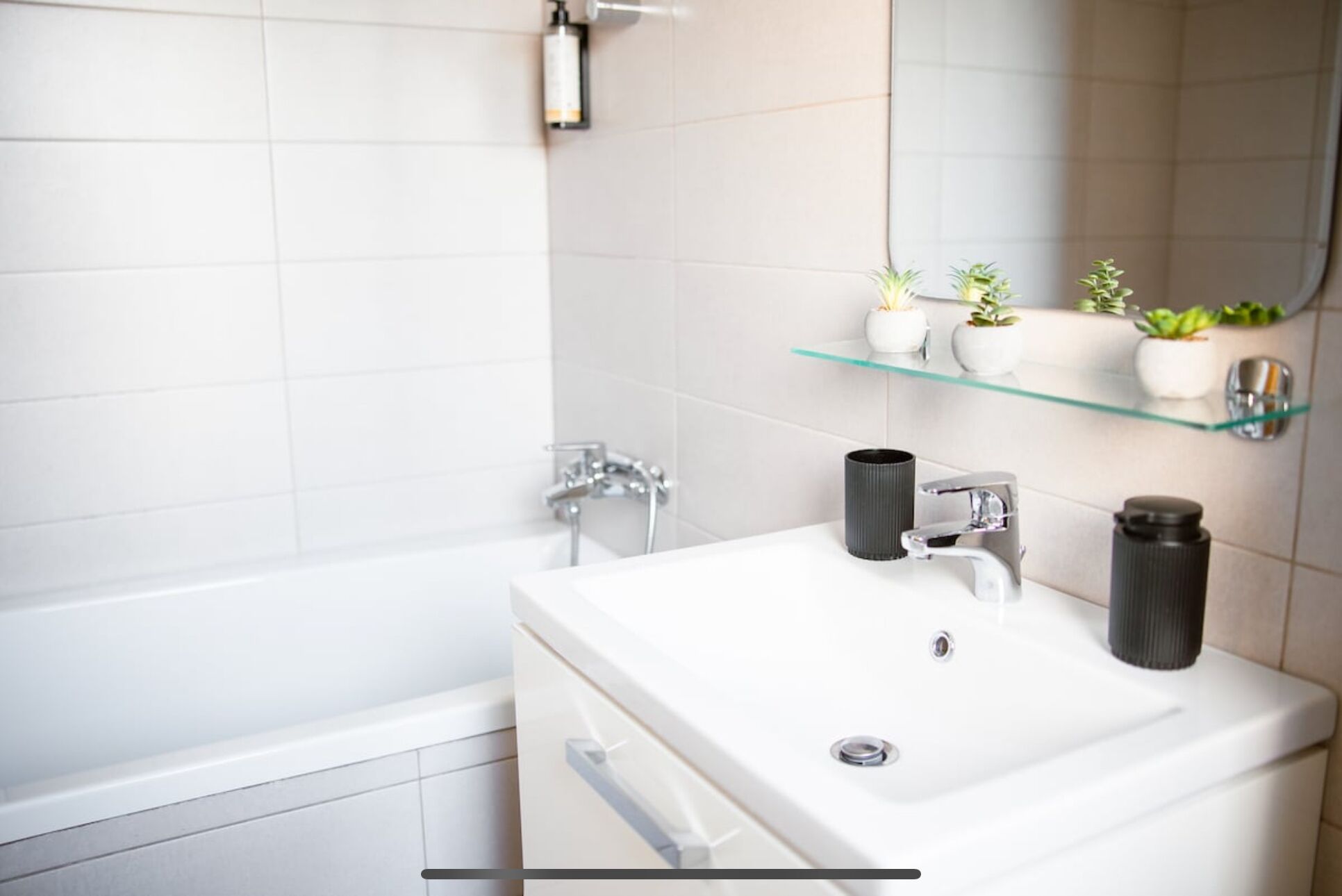 Ein modernes Badezimmer mit einem weißen Waschbecken, zwei Wasserhähnen, einem Spiegel und einer Glasablage mit Blumentöpfen.