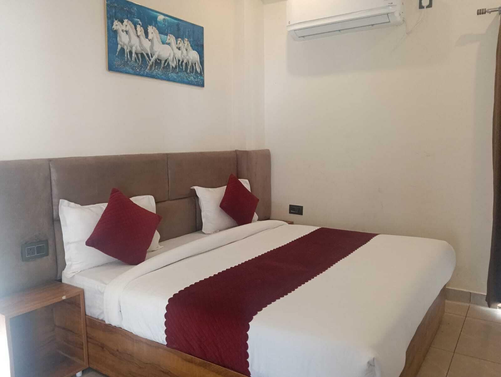 A bedroom with a bed, two pillows, a nightstand, a wooden headboard, a wall-mounted air conditioner, and a painting of horses.