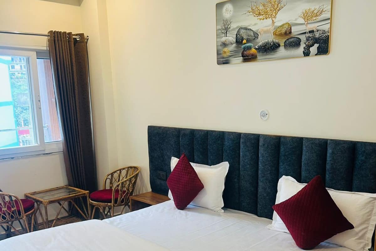 A hotel room with a large bed, two red pillows, a headboard, a window with curtains, a small table, and a chair.