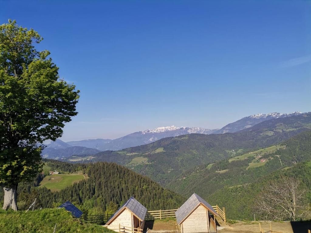Un paisaje montañoso con dos cabañas de madera, una con techo inclinado y otra con techo a dos aguas, rodeadas de abundante vegetación y picos nevados a lo lejos.