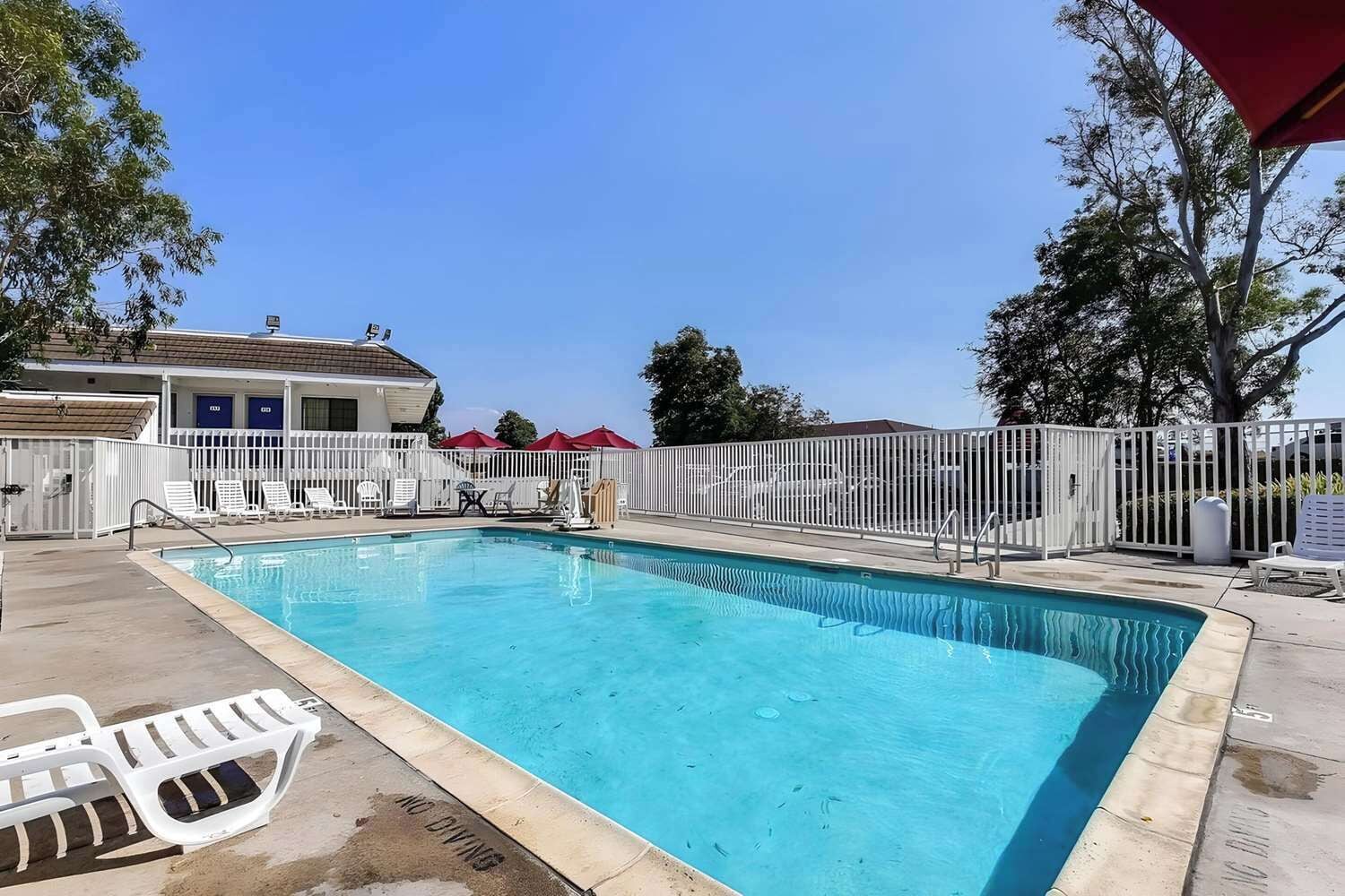 A swimming pool with a white fence, lounge chairs, and a building with a blue door in the background.