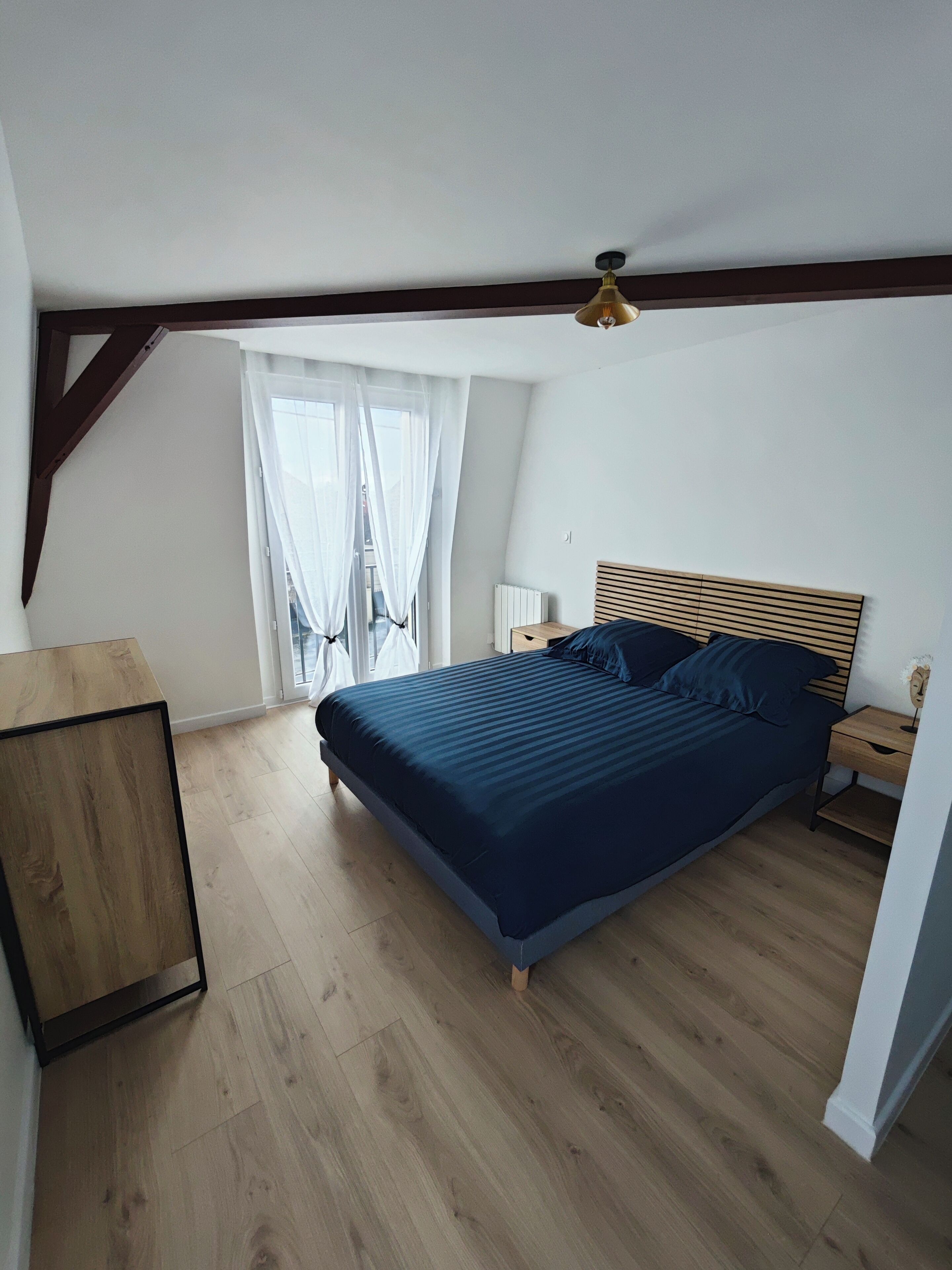 Une chambre à coucher avec un lit en bois, une table de chevet, un bureau en bois et une fenêtre ornée de rideaux légers.