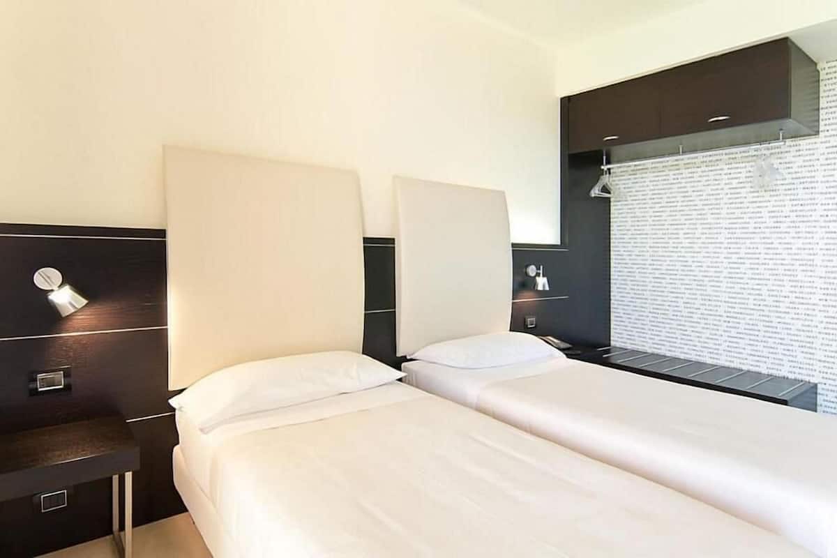 Habitación de hotel con dos camas, un cabecero oscuro, una mesita de noche y una ventana con persianas.