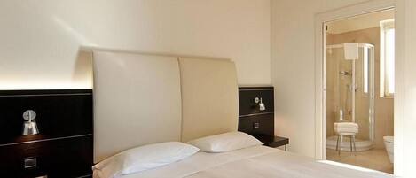 Habitación de hotel con cama, mesita de noche con teléfono, espejo y puerta que da a un baño con ducha e inodoro.