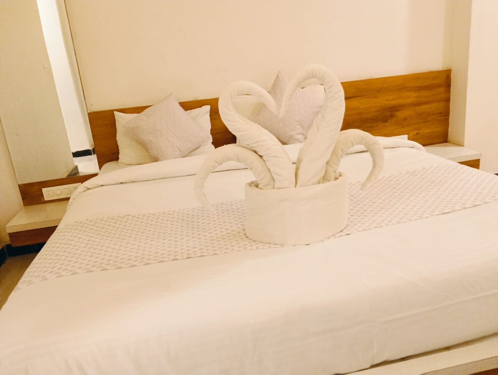 Een netjes opgemaakt bed met een handdoekarrangement in de vorm van een hart in een witte vaas, een houten hoofdbord en een wit kussen.