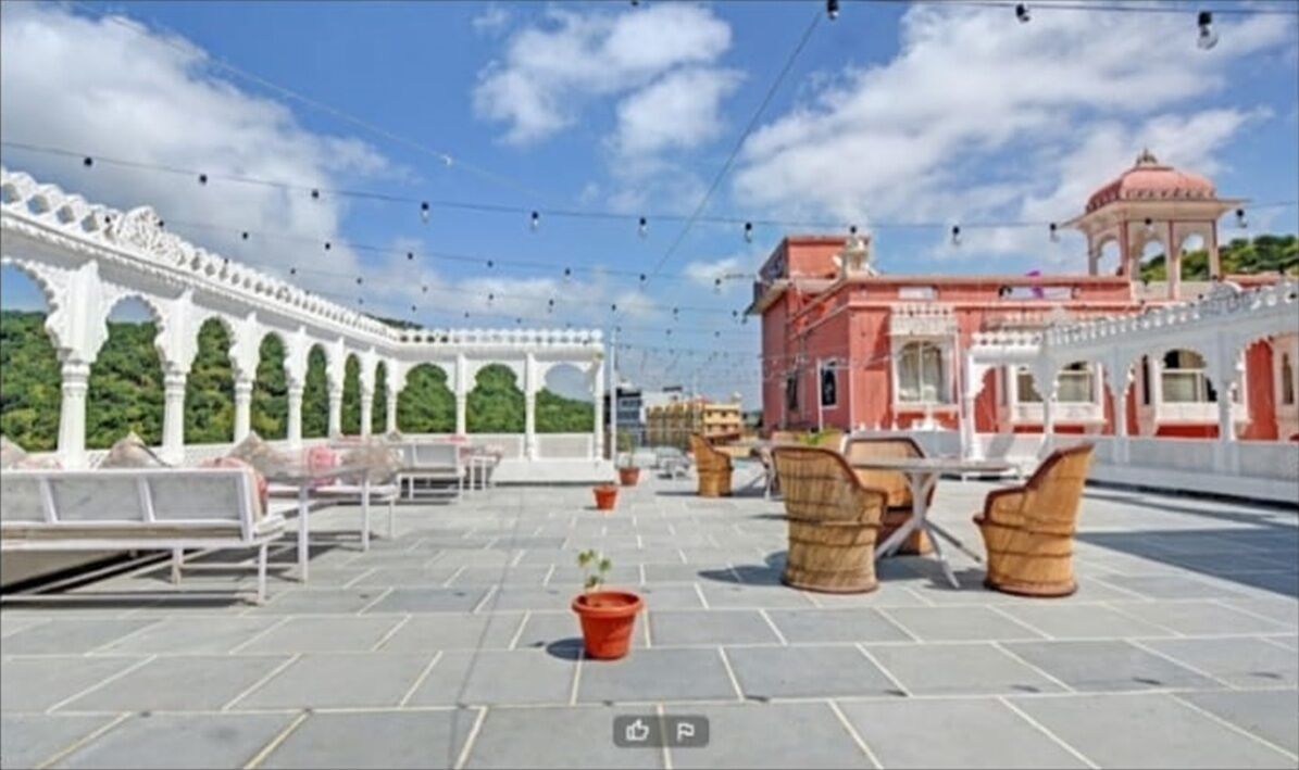 Terraço no topo com áreas de estar, uma pequena planta em vaso e vista para um edifício rosa com arcos.