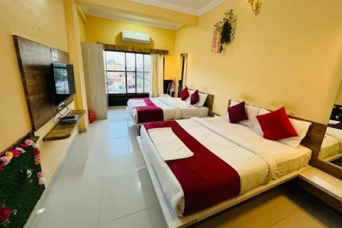 A hotel room with three beds, each with red and white bedding, a flat-screen TV, and a window with curtains.