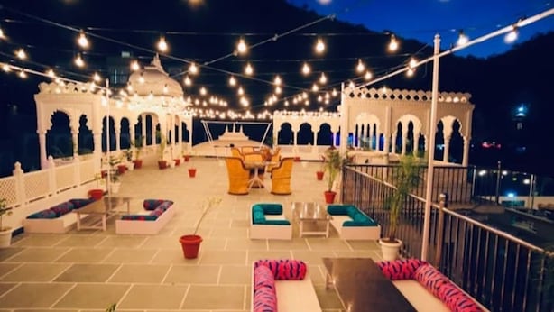 An outdoor seating area with cushioned benches and tables, string lights overhead, and a view of the cityscape at night. - Leopard Valley Resort (Kumbhalgarh)