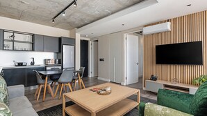 A modern living room with a wooden coffee table, a green sofa, a TV mounted on a wooden wall, and a kitchen area with grey cabinets and a black countertop. - Harbourline Haven | Signature #304 | De Waterkant (Cape Town)