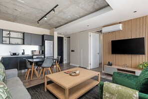 Ein modernes Wohnzimmer mit Couchtisch aus Holz, einem grünen Sofa, einem an einer Holzwand montierten Fernseher und einer Küchenzeile mit grauen Schränken und einer schwarzen Arbeitsplatte.