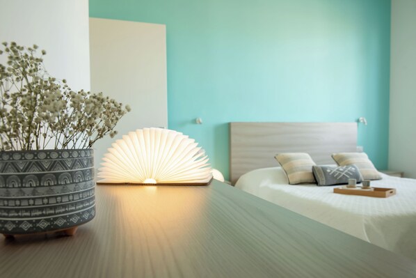 Ein ordentlich bezogenes Bett mit Kissen und Tablett, ein Blumentopf mit Blumen sowie ein Buch und eine fächerförmige Lampe auf einem Holztisch.