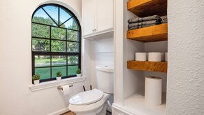 A bathroom with a toilet, a window with a view of greenery, and shelves with towels.