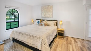 A bedroom with a large bed, two bedside lamps, a window with a view of a green lawn, and a wall-mounted abstract art piece.