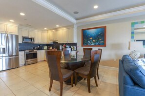 A modern kitchen with white cabinets, stainless steel appliances, and a dining area with a glass table and wicker chairs. - Ocean View, Pool, Elevator, Bed Linens/Bath Towels (Wildwood Crest)