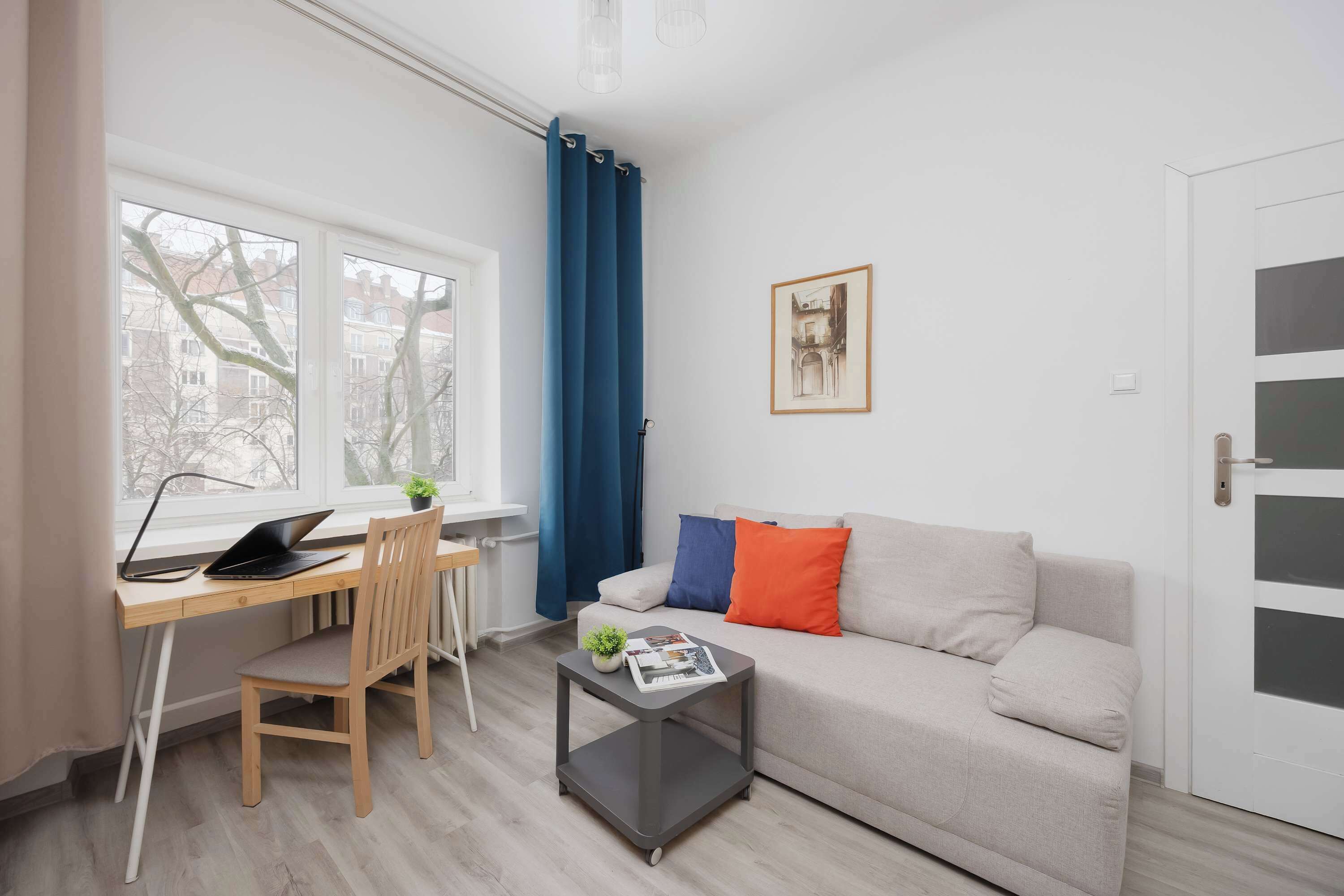 Ein modernes Wohnzimmer mit einem Holztisch, einem grauen Couchtisch, einem beigefarbenen Sofa und einem Fenster mit blauen Vorhängen.