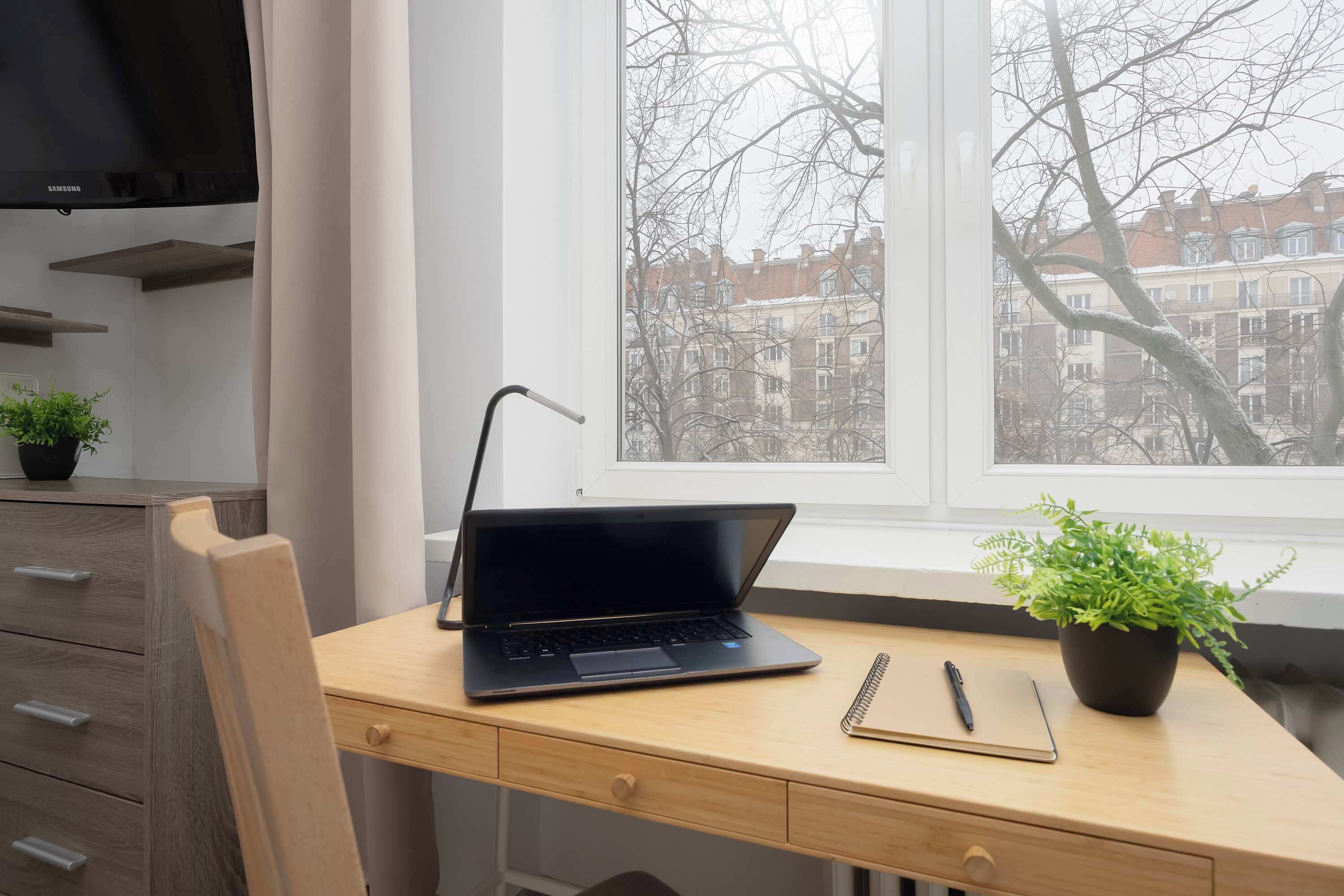 A laptop on a wooden desk by a window with a view of buildings.