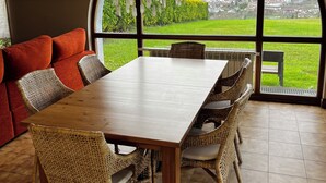 Ein Essbereich mit Holztisch und Korbstühlen, großen Bogenfenstern mit Bergblick und einem roten Sofa.
