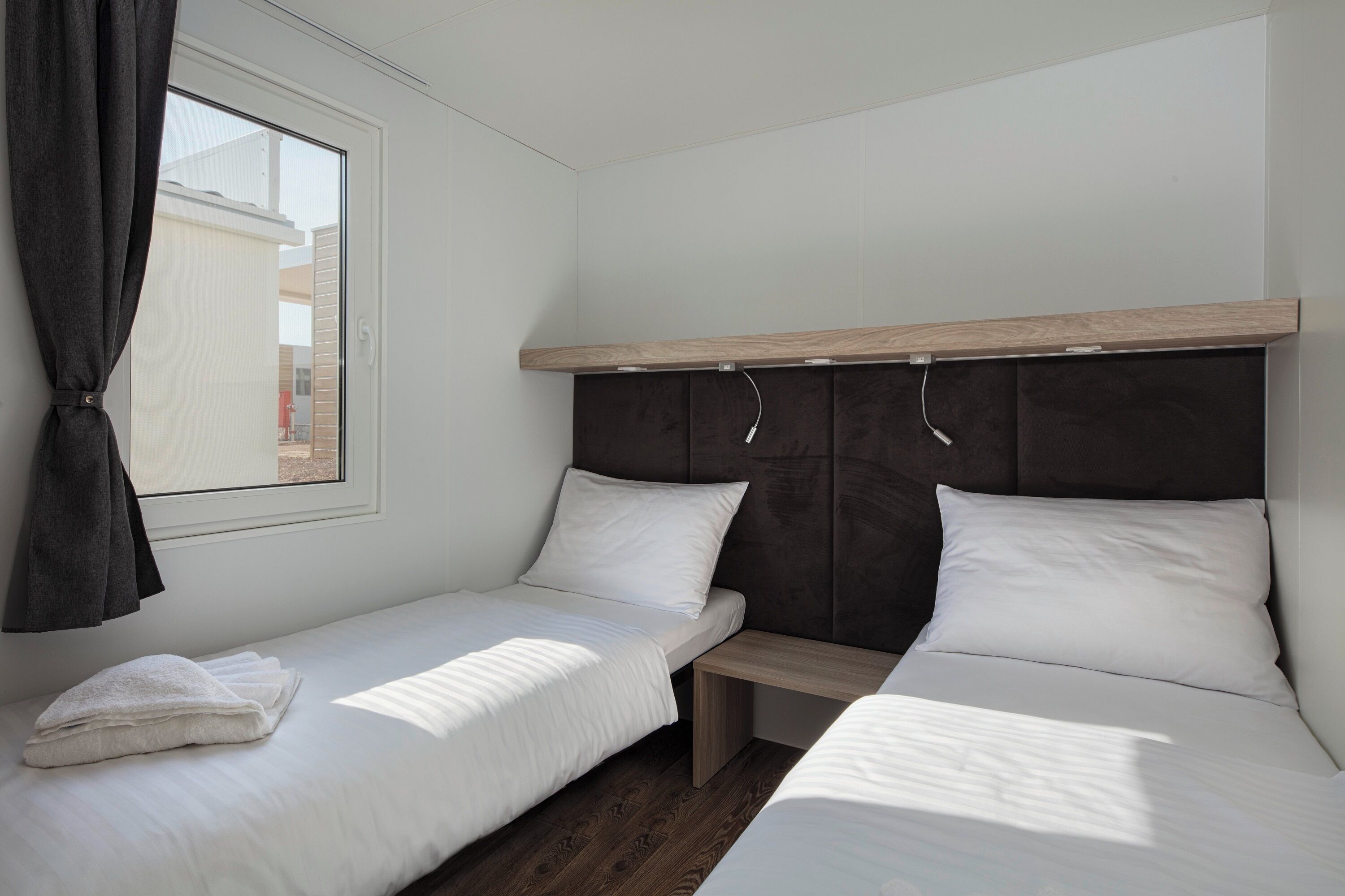 A bedroom with two single beds, a wooden headboard, a window with curtains, and a small bedside table.