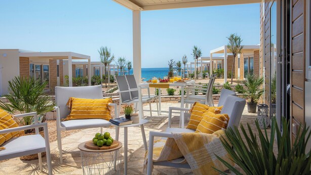 Een overdekt terras met witte stoelen en gele kussens, met uitzicht op een strand en de zee.