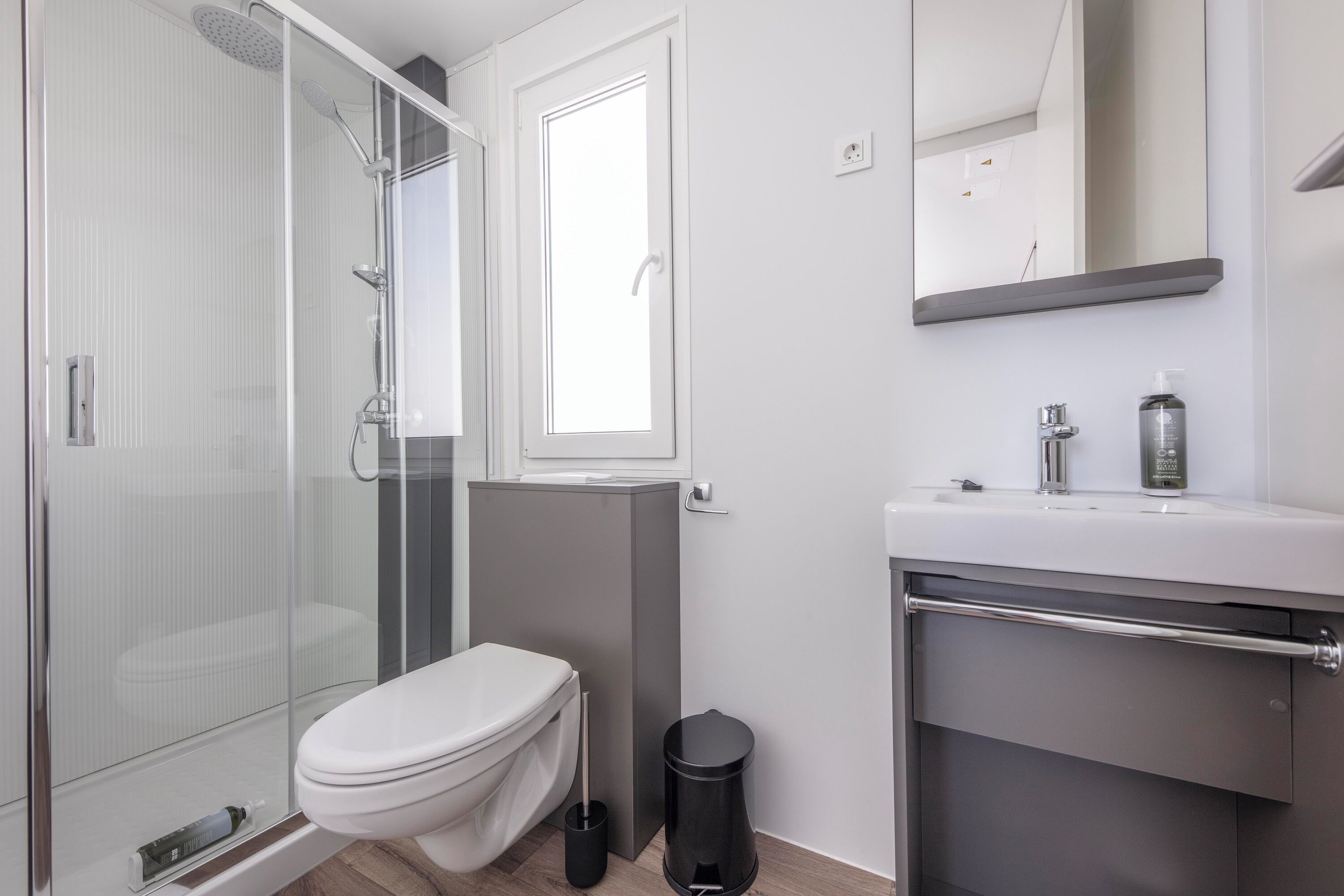Une salle de bain moderne avec une douche à parois de verre, des toilettes, une poubelle et un lavabo avec un miroir au-dessus.