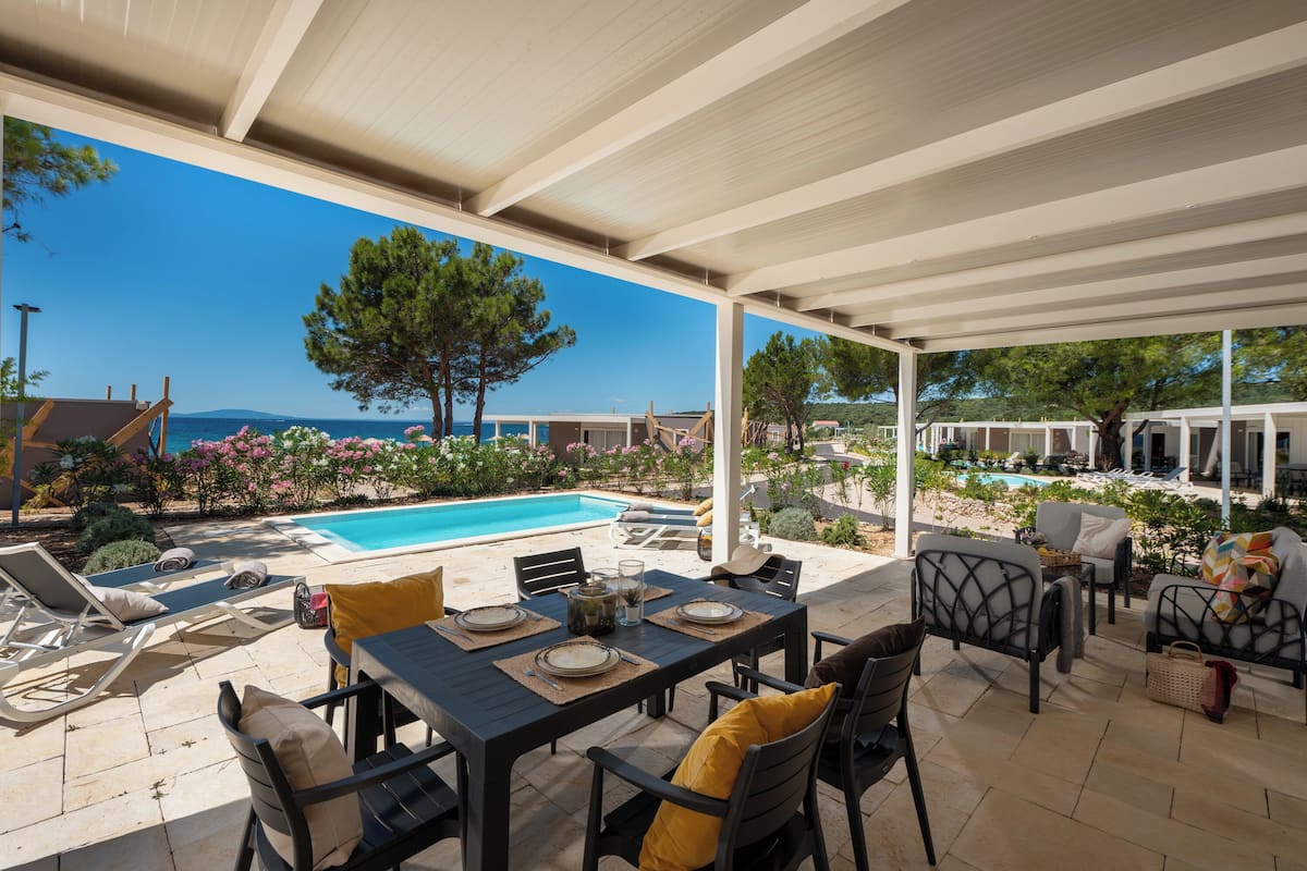 Um pátio externo com piscina, mesa de jantar e cadeiras, com vista para um céu azul límpido e o mar.
