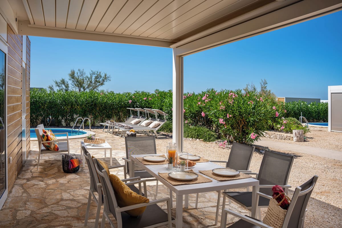 Uma área de refeições coberta ao ar livre com mesas e cadeiras, piscina e um jardim com flores cor-de-rosa.
