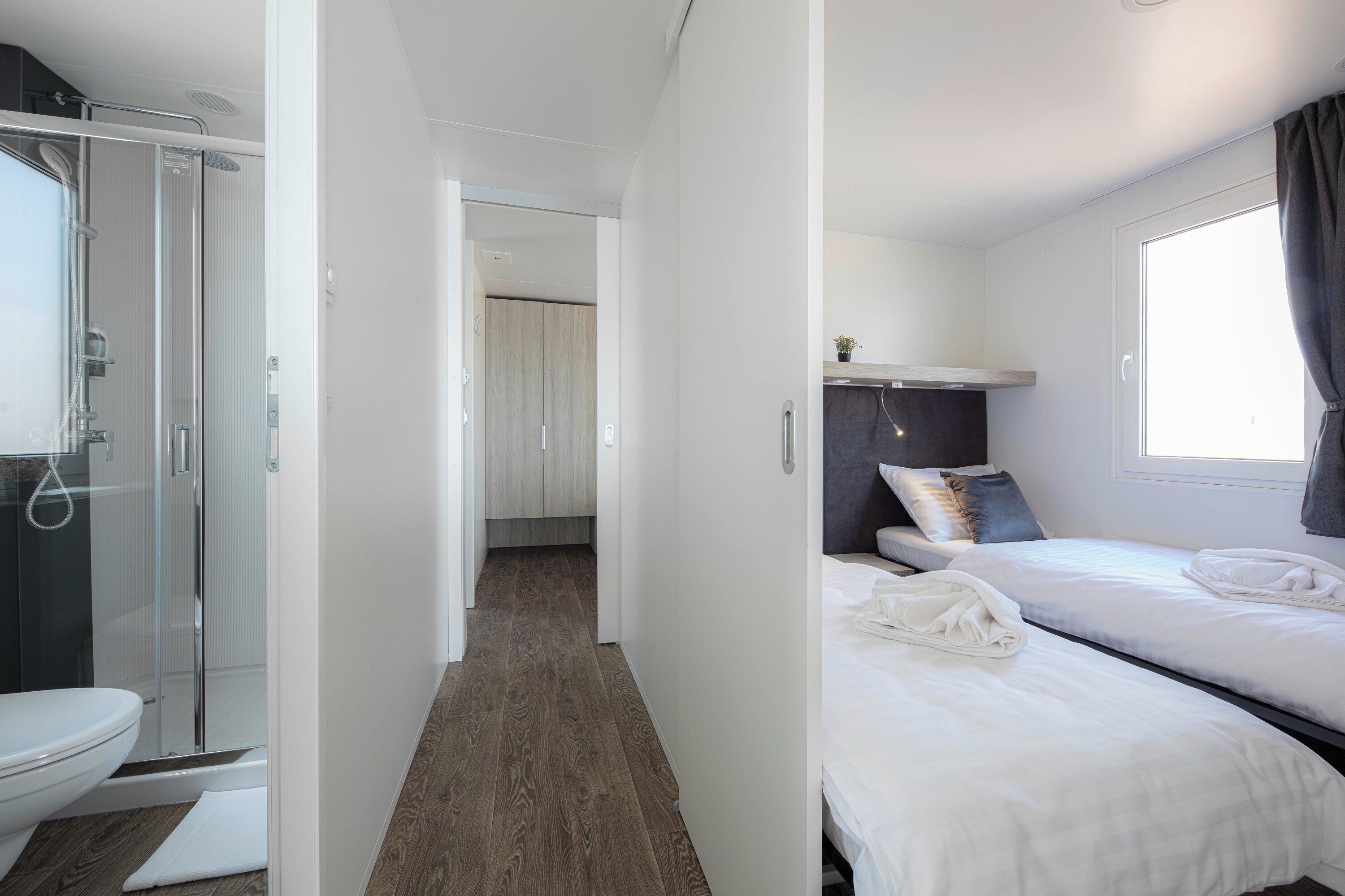A modern hotel room with a shower, a separate toilet, a bed with white linens, and a small window with curtains.