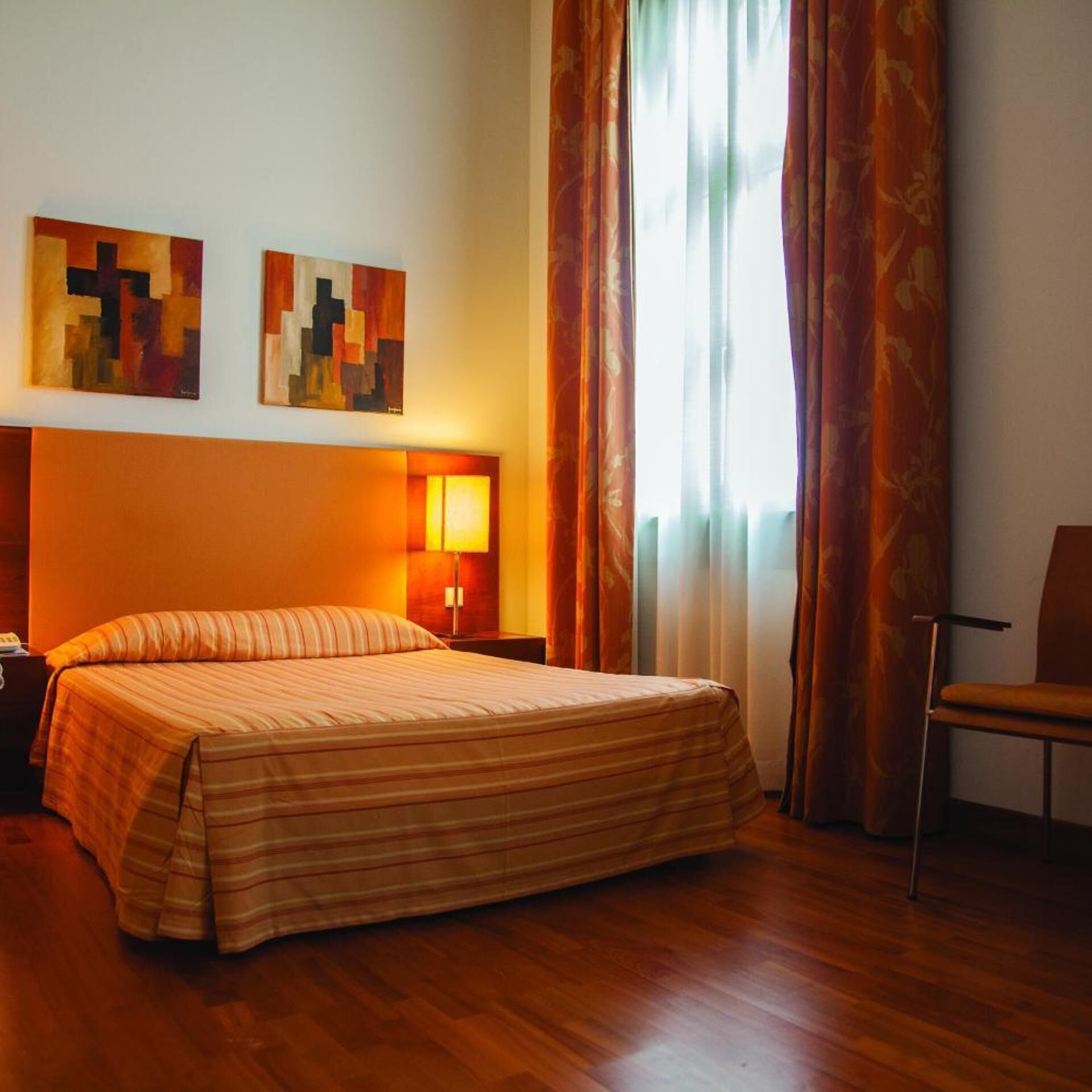 Habitación de hotel con cama, mesita de noche, lámpara, silla y dos cuadros en la pared.
