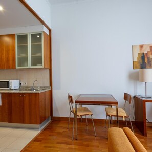 A compact kitchen with a microwave, sink, and wooden cabinets. There is a dining table with chairs and a sofa in the living area. - Hotel Apartamento Geres Ribeiro (Terras de Bouro)