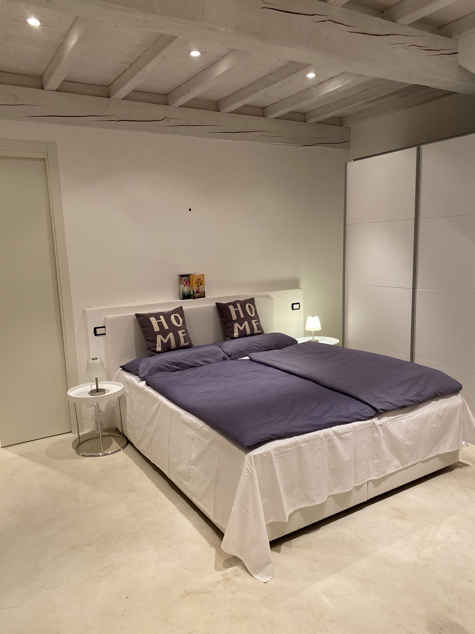 A bedroom with a bed, white walls, a ceiling with exposed beams, a bedside table, and a nightstand.