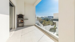 Uma varanda com chão de azulejos, pequena lareira de tijolos e guarda-corpo de vidro com vista para uma área residencial.
