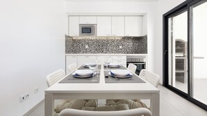 Uma área de jantar moderna com uma mesa branca posta para quatro pessoas, uma cozinha ao fundo com armários brancos e painel atrás do fogão, e uma grande janela com vista para a cidade.
