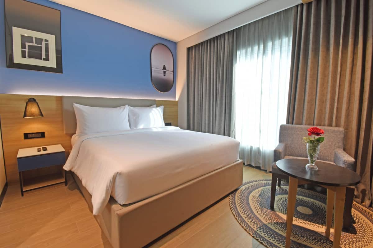 A hotel room with a large bed, a bedside table, a chair, a vase with a flower, a wall-mounted picture, and a window with curtains.